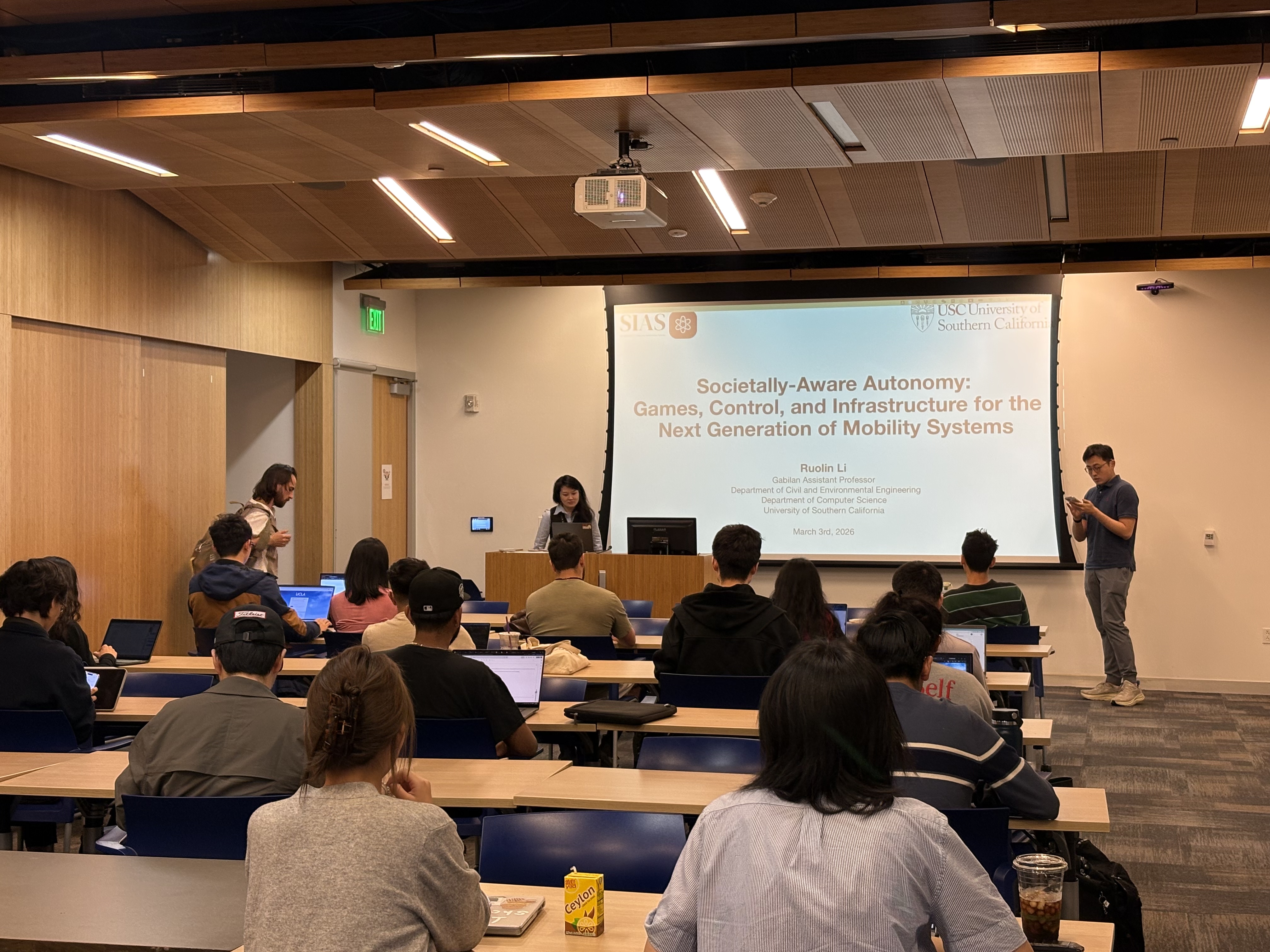 Ruolin presenting her societally-aware autonomy talk to an audience at UCLA, with the talk title projected behind her.
